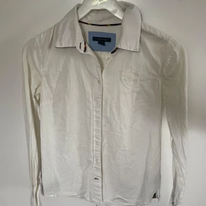 Vit skjorta från Tommy Hilfiger - Klassisk vit skjorta från Tommy Hilfiger med lång ärm och diskret broderad logga på bröstet. Skjortan har knappar framtill och en stilren krage. Perfekt för en clean och snygg look.