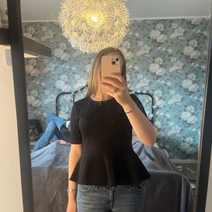 Svart peplumtop från H&M - Säljer en svart peplumtop från H&M med korta ärmar och rund halsringning. Toppen har en strukturerad yta och markerad midja som ger en snygg siluett. Perfekt att styla med jeans eller kjol.