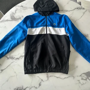 Blå och svart vindjacka från Lacoste - Snygg vindjacka från Lacoste Sport i blått, svart och vitt. Jackan har huva, dragkedja framtill och två fickor. Regular fit och tillverkad i lätt syntetmaterial. Klassisk Lacoste-logga på bröstet och elastisk nederkant för skön passform.