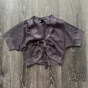 Grå bolerotopp med spets och knyt - Supersöt grå bolerotopp med korta ärmar och fina spetsdetaljer längs kanterna. Toppen är croppad och har knyt framtill för en extra gullig look. Perfekt att slänga över ett linne eller klänning för en trendig layering eller som den är med ett par jeans 