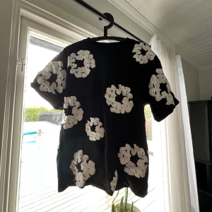 Svart t-shirt från Denim Tears - Svart t-shirt från Denim Tears med stora vita blommiga mönster över hela plagget. Klassisk passform med korta ärmar och rund hals. Tillverkad i mjuk bomull, perfekt för dig som vill sticka ut med en unik och trendig stil. Stl S men sitter Som M! 