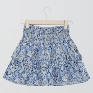 Blåvit volangkjol från Gina Tricot - Supersöt kort kjol från Gina Tricot med blått och vitt blommönster. Kjolen har smock i midjan och flera lager volanger som ger en flowy känsla. Perfekt för varma dagar och lätt att matcha med en enkel topp. Tyvärr har några trådar i resåren gått upp därav priset!