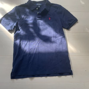 Marinblå pikétröja från Ralph Lauren - Snygg marinblå pikétröja från Polo Ralph Lauren med klassisk krage, två knappar och röd broderad logga på bröstet. Tillverkad i mjuk bomull och har korta ärmar. Perfekt för en clean och stilren look.