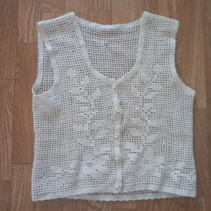 Vit virkad väst med blommönster - Superfin virkad väst i vitt med blommönster och knappar framtill. Västen har en luftig, nätliknande struktur och v-ringad hals. Perfekt att slänga över en t-shirt eller klänning för en chill och trendig look.