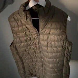 Olivgrön quiltad dunväst - Säljer en olivgrön quiltad dunväst med dragkedja framtill och hög krage. Jag är 1,75. Västen har en klassisk, lätt vadderad look och är perfekt att slänga över en hoodie eller tröja. Enkel och stilren design utan synliga loggor eller mönster. Helt ny 