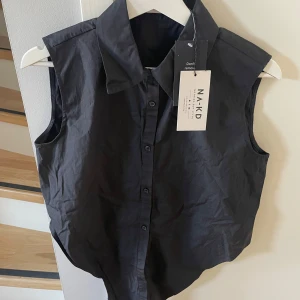 Svart ärmlös skjorta från NA-KD - Snygg svart ärmlös skjorta från NA-KD med knappar framtill. Perfekt för en stilren look. Skjortan har en klassisk krage och är designad för att knytas framtill.