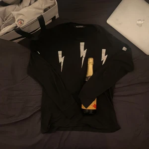 Svart tröja från Neil Barrett - Säljer en svart långärmad tröja från Neil Barrett med vita blixtmotiv på framsidan. Perfekt för en stilren och modern look. Passar bra till både jeans och chinos. Nypris 3000