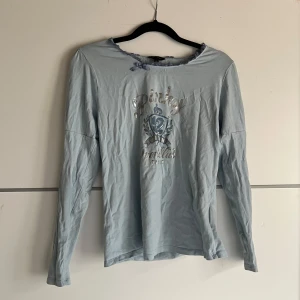 Ljusblå långärmad topp från Byer California - Säljer en ljusblå långärmad topp från Byer California med ett tryck framtill. Toppen har en rund halsringning med spetsdetaljer och är perfekt för en avslappnad stil. Passar bra till jeans eller kjol för en casual look.