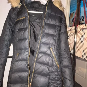 Svart dunjacka med pälsdetaljer från Rockandblue - Snygg svart dunjacka från Rockandblue med pälsdetaljer på huvan. Jackan har dragkedjor i guld och flera praktiska fickor. Perfekt för kyliga dagar med sin quiltade design och långa ärmar, säljs oxå pågrund av flykt. Pris kan diskuteras, 