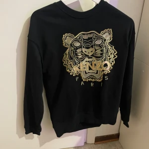 Svart sweatshirt från Kenzo - Säljer en svart sweatshirt från Kenzo med ett stort broderat tigerhuvud i guld på framsidan. Tröjan har långa ärmar och en rund halsringning. Perfekt för en stilren och trendig look.
