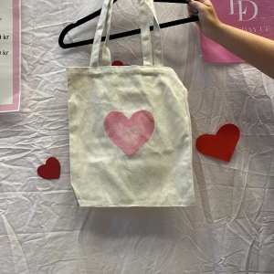 Vit shoppingväska med rosa hjärta - Vi är ett UF företag som säljer tygväskor med handmålade tryck på framsidan. Perfekt för att bära med sig det nödvändigaste med stil.