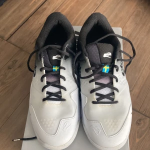 Vita och svarta sneakers från Salming - Snygga vita och svarta sneakers från Salming med svensk flagga på sidan. Skorna har svarta snören och en stilren design med Salming-logga. Perfekta för sportiga aktiviteter eller vardagsbruk. Storlek 40 2/3