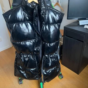 Svart dunväst från H&M Divided - Snygg svart dunväst från H&M Divided med glansig yta och dragkedja framtill. Perfekt för att hålla värmen med stil. Västen har en modern look och passar bra till olika outfits.