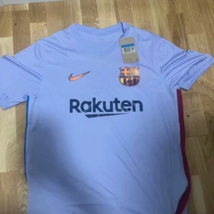Lila fotbollströja från Nike - Säljer en lila fotbollströja från Nike med korta ärmar. Tröjan har en V-ringning och klubbens emblem på bröstet. Den har även sponsorlogga i svart och en orange Nike-logga. Perfekt för fotbollsfans!