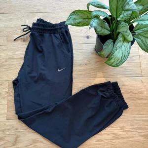 Svarta mjukisbyxor från Nike - Säljer ett par svarta mjukisbyxor från Nike med elastisk midja och snörning. Byxorna har en liten Nike-logga på sidan och är perfekta för en avslappnad stil. De är tillverkade i ett mjukt och bekvämt material.