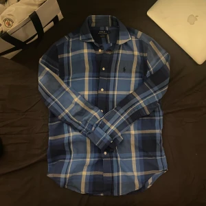 Blå rutig skjorta från Ralph Lauren - Snygg blå rutig skjorta från Ralph Lauren med klassisk krage och knappar framtill. Skjortan har långa ärmar och ett broderat logotypmärke på bröstet. Perfekt för en stilren look.