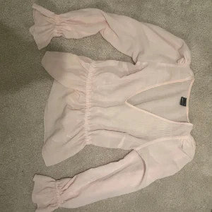 Ljusrosa blus från Gina - Säljer en söt ljusrosa blus från Gina med v-ringning och långa ärmar. Blusen har en luftig passform och elastiska detaljer vid ärmsluten. Perfekt för en stilren look!