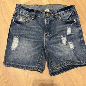Blå jeansshorts från Uniq Young - Säljer ett par coola blå jeansshorts från Uniq Young med slitna detaljer. Shortsen har en klassisk femficksdesign och knappgylf. Perfekta för en avslappnad stil.