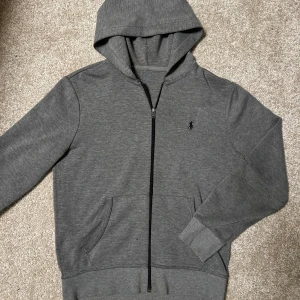 Ralph Lauren zip hoodie - Säljer en stilren mörkgrå zip hoodie från Ralph Lauren med dragkedja och huva. Tröjan har långa ärmar och en liten logotyp på bröstet. Perfekt för en avslappnad look.