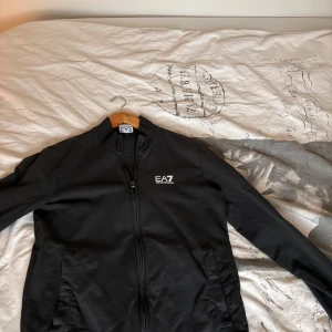 Svart jacka från Emporio Armani - Säljer en stilren marinblå ish tröja från Emporio Armani EA7. Tröjan har en dragkedja framtill och är långärmad. Perfekt för en sportig look med sin enkla design och diskreta logga på bröstet.