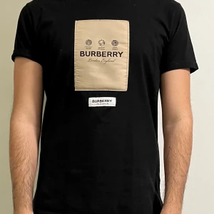 Svart t-shirt från Burberry - Snygg svart t-shirt från Burberry med en beige patch på bröstet där märkesnamnet och 'London, England' är tryckt.  Passformen är M/L, ÄKTA.