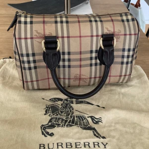 Rutig handväska från Burberry, äkta - Säljer en klassisk rutig handväska från Burberry i beige, svart och röd. Väskan har svarta handtag och en dragkedja upptill. Perfekt för att ge en stilren touch till din outfit.