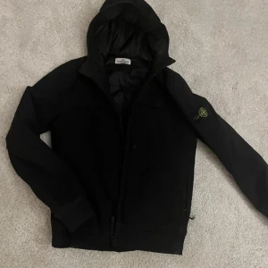 Svart jacka med huva från Stone Island - Snygg svart jacka från Stone Island med huva och dragkedja. Jackan har en ikonisk logga på ärmen och är perfekt för kyligare dagar. Den har en stilren design och är långärmad.