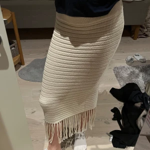 Beige stickad kjol med fransar - Snygg beige stickad kjol med fransar längst ner. Kjolen har en tight passform och är perfekt för en stilren look. Den är knälång och ger en bohemisk touch till din outfit.