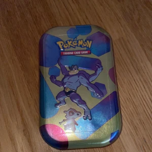 Pokémon Trading Card Game Mini Tin 151 card madr in USA - Säljer en Pokémon Trading Card Game Mini Tin med motiv av Machamp och Cubone. Perfekt för att förvara dina kort eller som samlarobjekt. Tinnet har en färgglad design med holografiska detaljer och innehåller booster packs från Scarlet & Violet-151 serien.