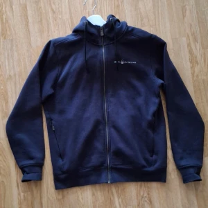 Mörkblå hoodie från Sail Racing - Säljer en mörkblå hoodie från Sail Racing med dragkedja och hög krage. Den har långa ärmar och en justerbar huva med snörning. Perfekt för en avslappnad stil. Använd ett fåtal gånger. Hör gärna av dig om du har några frågor! 