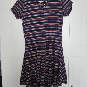 Randig klänning från Guess - Säljer en randig klänning från Guess i mörkblått med inslag av lila, rosa och orange. Klänningen har korta ärmar och en snygg snörning framtill. 