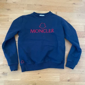Mörkblå sweatshirt från Moncler - Säljer en snygg mörkblå sweatshirt från Moncler med röd broderad logga på framsidan. Tröjan har långa ärmar och ribbade muddar vid ärmslut och nederkant. Perfekt för en stilren look!