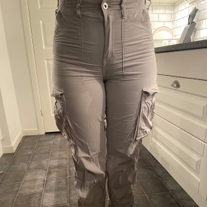 Beige cargobyxor från Bershka - Snygga beige cargobyxor från Bershka med hög midja och flera praktiska fickor. Perfekta för en avslappnad stil med en touch av funktionalitet. Byxorna har en klassisk cargostil med en modern twist. Byxknappen på höger bakficka saknas.