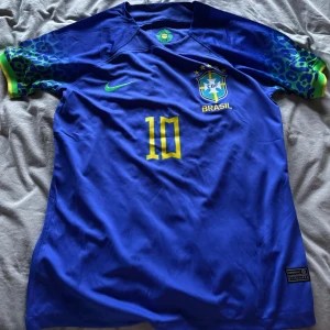 Blå Brasilien fotbollströja - Neymar  - Säljer en blå Brasilien fotbollströja  med nummer 10 och texten 'Neymar Jr' på ryggen. Tröjan har korta ärmar med ett unikt mönster i grönt och gult. Perfekt för fotbollsfans som gillar Brasilien. Tröjan är perfekt nu till sommaren och är aldrig använd. 