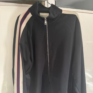 Svart jacka från Gucci - Snygg svart jacka från Gucci med dragkedja och långa ärmar. Jackan har vita och röda ränder längs ärmarna som ger en sportig look. Perfekt för dig som vill ha stil och komfort i ett.