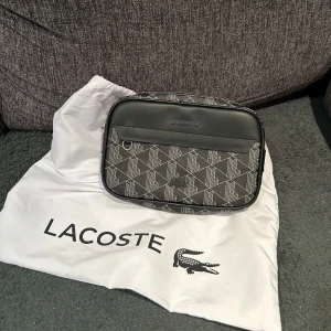 Svart axelväska från Lacoste - Snygg svart axelväska från Lacoste med ett snyggt mönster. Väskan har en dragkedja upptill och en justerbar axelrem. Perfekt för att bära dina nödvändigheter med stil och har ett stort utrymme också. Finns möjlighet för byte också!