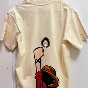 Beige t-shirt med One Piece-motiv - Säljer en beige t-shirt från Uniqlo med ett coolt One Piece-tryck. Framsidan har en stor logga med en hatt och dödskalle, medan baksidan visar en karaktär med uppsträckt arm. Perfekt för anime-fans!