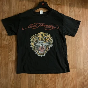 Svart t-shirt med tigertryck från Ed Hardy - Säljer en svart t-shirt från Ed Hardy med ett stort tigertryck på framsidan och röd logotyp både fram och bak. Perfekt för dig som gillar Ed Hardy för att den är äkta och att den är på bra skick  