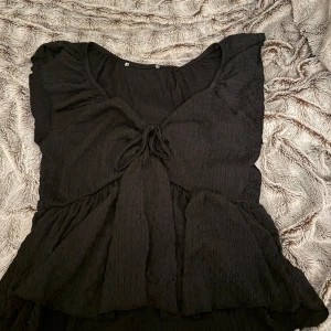 Svart blus med volangdetaljer - Säljer en svart söt blus med volangdetaljer och knytning framtill. Blusen har en lös passform och korta armar. Säljer för att inte använder längre❤️