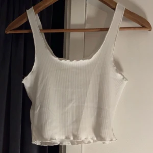 Vit ribbad linnetopp - Säljer en stilren vit ribbad linnetopp med breda axelband. Toppen har en enkel och klassisk design som passar perfekt till sommaren. Den är croppad och har en lätt vågig kant. Materialet är även väldigt svalkande vilket är perfekt till sommaren. Linnet är endast testad och finns även i svart som jag säljer🥰