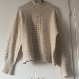 Beige tröja med pärlor från H&M - Säljer en stilren beige tröja från H&M dekorerad med pärlor längs ärmarna och framtill. Tröjan har en hög krage och långa ärmar, perfekt för en elegant look. Passar utmärkt till både vardag och speciella tillfällen.