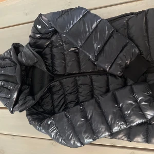 Svart dunjacka från Peak Performance - Säljer en svart dunjacka från Peak Performance. Jackan har en glansig yta, perfekt för kyligare men också soligare dagar.