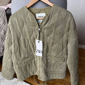 Olivgrön quiltad jacka från Zara - Säljer en olivgrön quiltad jacka från Zara med dragkedja framtill. Jackan har en unik vågig quiltning och två fickor. Den är aldrig använd med lappen kvar i storlek XS
