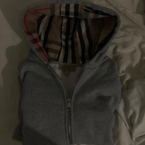 Grå hoodie från Burberry - Säljer en stilren grå hoodie från Burberry med dragkedja och klassiskt rutigt mönster i huvan. Perfekt för en avslappnad och trendig look. Hoodien har långa ärmar och en broderad logga på bröstet. Nypris 7900kr