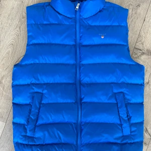 Blå dunväst från GANT - 🔵 Säljer en snygg blå dunväst från GANT med dragkedja framtill. Knappt använd och i fint skick! 🔵