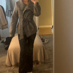 Grå kappa med knytband - Säljer en stilren grå kappa med knytband i midjan. Jackan har ett fiskbensmönster och långa ärmar, perfekt nu till våren. Jättefin passform