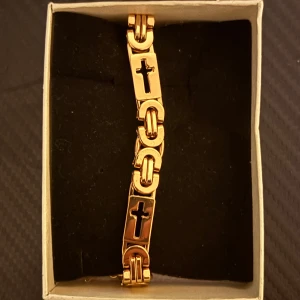 Guldarmband med korsdetaljer - Elegant guldarmband med korsdetaljer. Perfekt för att ge en stilfull touch till din outfit. Armbandet har en klassisk design med flera korslänkar. detta armband är tillverkat med rostfritt stål bredd 9cm, Längd 23cm