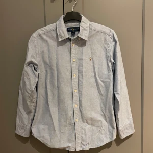 Ljusblå skjorta från Ralph Lauren - Säljer en stilren ljusblå skjorta från Ralph Lauren med klassisk krage och broderad logotyp på bröstet. Skjortan har långa ärmar och knappar framtill. Väldigt bra skick. Stl 14. 