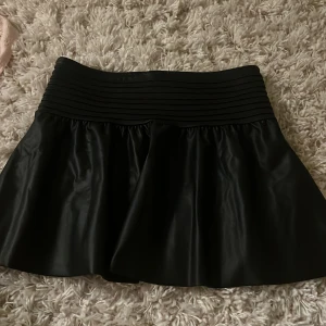 Svart kjol från Gina Tricot - Snygg svart kjol från Gina Tricot med en bred ribbad midja som ger en bekväm passform. Kjolen har en glansig finish och är perfekt för att skapa en stilren look. Passar utmärkt till både vardag och festliga tillfällen.