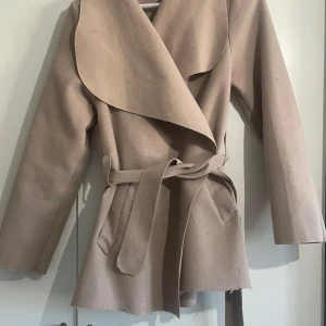 Beige kappa med bälte - Säljer en stilren beige kappa med bälte och långa ärmar. Den har en omlott-design och två fickor framtill. Perfekt för höst och vår! Den är avklippta till en kort kappa men inget som stör utseendet